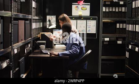 I partner della polizia interrazziale lavorano come investigatori privati, esaminando le prove nella sala d'archivio alla ricerca di indizi su un crimine. L'ispettore donna caucasico serio lavora con investigatore maschio concentrato sui fascicoli dei casi. Foto Stock