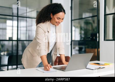 Una donna d'affari molto affollata, brasiliana o ispanica dai capelli ricci, ceo dell'azienda, responsabile del reclutamento, sta vicino a una scrivania in un ufficio moderno, lavora su un notebook su un progetto, sorridendo Foto Stock