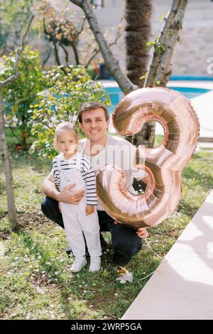 Papà sorridente che abbraccia una bambina che accovacciata in giardino con il numero 3 gonfiabile a corda Foto Stock