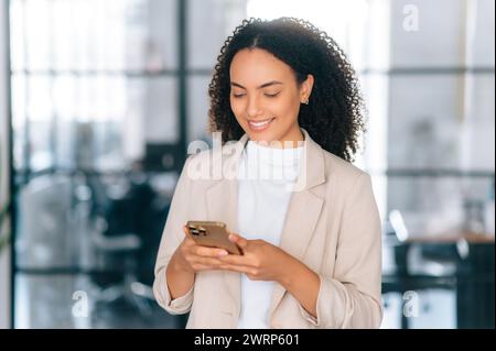 Uso di gadget. Splendida donna brasiliana o ispanica elegante, manager aziendale, segretaria, si trova in un ufficio moderno, utilizzando lo smartphone per chattare con il cliente o il partner, scrivendo e sorridendo Foto Stock