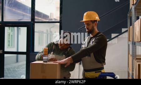 Dolly fuori dal capo della logistica nel magazzino supervisionando il tirocinante che sigilla i pacchi di cartone. Il personale del luogo di stoccaggio fissa correttamente le merci per evitare danni durante la spedizione Foto Stock