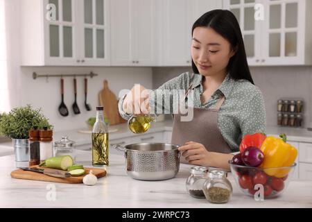 Processo di cottura. Bella donna che versa olio dalla bottiglia alla pentola sul piano bianco in cucina Foto Stock