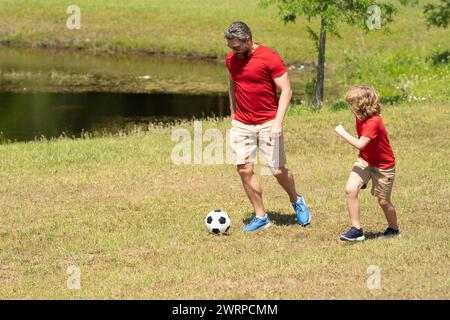 Padre con figlio che gioca a calcio sul campo. Padre e figlio giocano a calcio insieme nel parco estivo. Il figlio eccitatamente calcia la palla verso il padre Foto Stock