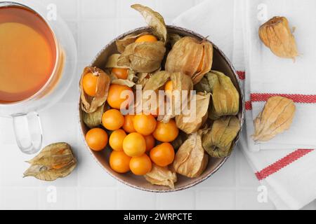 Frutti di physalis maturi con calici in ciotola e tazza di tè su un tavolo piastrellato bianco, disteso piatto Foto Stock