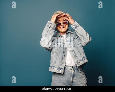 Donna matura e felice con i capelli grigi si trova in uno studio, indossando un abbigliamento alla moda in denim e occhiali da vista. Il suo sorriso giocoso e spensierato Foto Stock