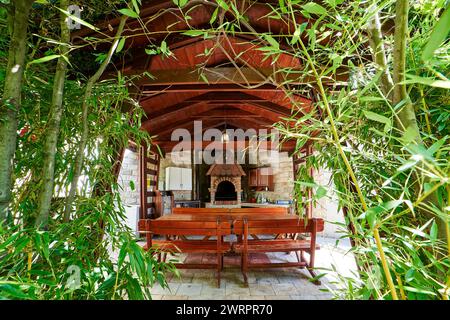 Vista attraverso i rami verdi del giardino su un gazebo in legno con piano cottura e armadi da cucina Foto Stock