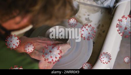 Immagine composita di cellule covid-19 che fluttuano contro il primo piano delle mani che lavorano su una ruota di ceramica Foto Stock