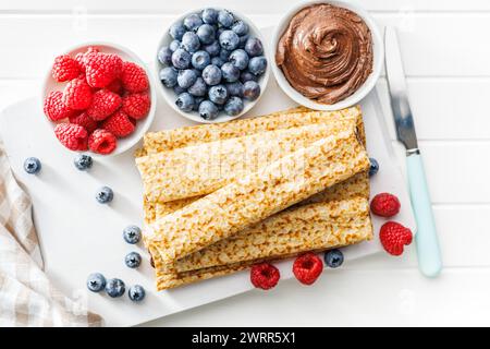 Crepe fresche ricoperte di frutti di bosco freschi servite su un piatto bianco accanto a ciotole di lamponi, mirtilli e spalmazione al cioccolato. Vista dall'alto. Foto Stock