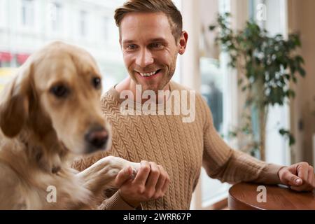 Primo piano ritratto di un bellissimo cane Golden retriever che dà la zampa a un uomo. Un ragazzo sorridente tiene il suo animale domestico mentre si siede in un bar Foto Stock