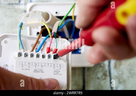 Elettricista che collega fili colorati ai terminali del termostato montato a parete per il riscaldamento a pavimento in edifici residenziali durante la ristrutturazione. Foto Stock