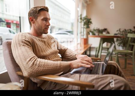 Concetto di persone lavoratrici e spazio pubblico. Giovane e bello, designer dell'interfaccia utente ux seduto con un laptop in un bar, bevendo un cappuccino, guardando lo schermo con Foto Stock