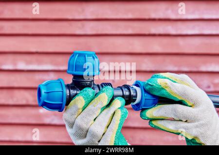 Raccordo a vite a T in PP per connettore morsetto acqua potabile, idraulico indossando guanti protettivi stringe il dado di serraggio, sullo sfondo di legno rosso wa Foto Stock