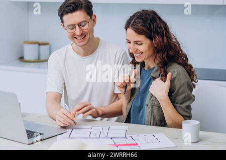 Una coppia sorridente sta lavorando a un nuovo progetto di appartamento, con un computer portatile e documenti con disegni della casa sparsi sul tavolo. Riscaldamento della famiglia. Foto Stock