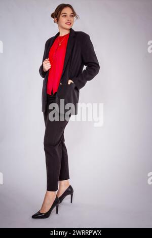 Una giovane donna bionda con un panino indossa un abito da pantaloni nero e una camicetta rossa. Combina tutto questo con eleganti tacchi alti. Lei pone con sicurezza in fron Foto Stock