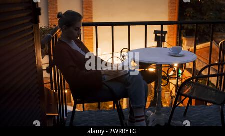 Una bella ragazza bionda legge un libro sul balcone della casa. Luce calda, notte accogliente. Romantico stile vintage e stile di vita Foto Stock