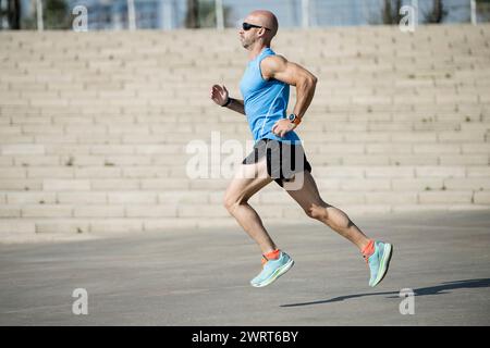Fai correre l'atleta in strada con una t-shirt blu e pantaloncini neri. Vista laterale di un atleta che fa jogging all'aperto. Foto Stock