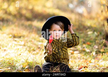Piccolo ragazzo sorpreso in un cappello a tegole seduto su un prato con foglie rosse autunnali in mano Foto Stock