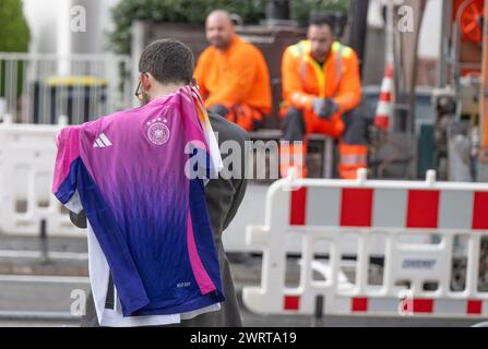 14 marzo 2024, Assia, Francoforte sul meno: I lavoratori della costruzione di strade guardano mentre un uomo cammina davanti al campus DFB, sede della federazione calcistica tedesca (DFB), indossando le nuove maglie ufficiali del campionato europeo della squadra DFB. L'allenatore nazionale Nagelsmann presenta la squadra del Campionato europeo in una conferenza stampa pomeridiana. La squadra della DFB giocherà contro la Francia a Lione il 23 marzo e contro i Paesi Bassi a Francoforte il 26 marzo. Foto: Boris Roessler/dpa Foto Stock