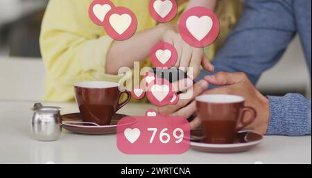Immagine delle icone del cuore che fluttuano sopra una coppia caucasica utilizzando lo smartphone e bevendo caffè Foto Stock