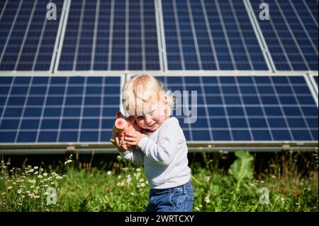 Bambino che cerca di scoprire quanti soldi ci sono nel suo salvadanaio sullo sfondo dei pannelli solari. Un bambino piccolo che impara a risparmiare denaro per il futuro. Concetto di risparmio di denaro, investimenti in energie rinnovabili. Foto Stock