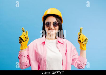 Donna con casco giallo e guanti che puntano e gesturano contro lo sfondo blu dello studio. Foto Stock