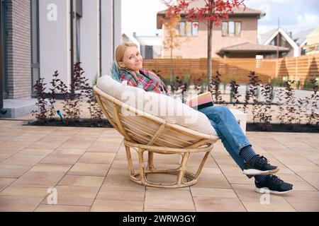 Donna sorridente avvolta in una coperta che riposa nel cortile della casa su una poltrona rotonda in vimini mentre legge un libro Foto Stock
