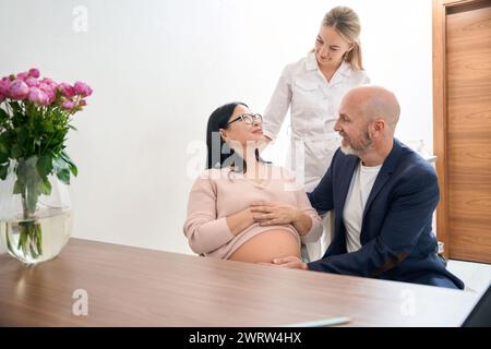 Attraente donna ginecologo saluto donna asiatica incinta e suo marito che è venuto a appuntamento medico il terzo trimestre di gravidanza, salute che Foto Stock