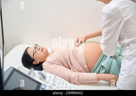 Ginecologo femminile che controlla l'altezza della pancia di donna incinta utilizzando il nastro di misurazione, controllo del feto che si sviluppa all'interno del grembo della donna, assistenza sanitaria Foto Stock