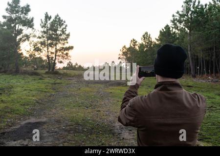 Vista posteriore di un uomo irriconoscibile in piedi in una giacca di pelliccia e in un cappello di lana nella foresta che scatta una foto con il suo telefono cellulare Foto Stock