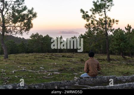 Vista posteriore di un uomo seduto nella foresta che guarda le isole Cies all'orizzonte al tramonto Foto Stock