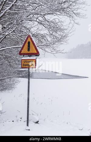 Un segno triangolare di avvertimento con un punto esclamativo si distingue su uno sfondo di bianco, che avverte di ghiaccio debole su un corpo d'acqua ghiacciato in Svezia Foto Stock