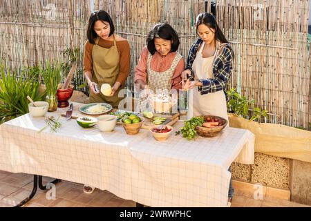 La madre del sud-est asiatico e le sue figlie si divertono a cucinare insieme una ricetta di cibo tailandese nel cortile Foto Stock