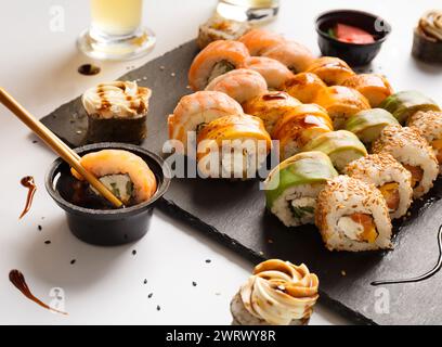 Un set di sushi con gamberi, salmone, avocado su un piatto di pietra nera, un rotolo è immerso in salsa di soia con bacchette cinesi. Foto Stock