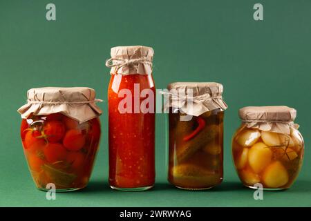 Verdure in scatola in vasetti di vetro chiusi su sfondo verde. Cetrioli sottaceto, pomodori, salsa al peperoncino, aglio, adjika. Spuntino piccante, cibo in scatola. Foto Stock