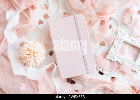 Notebook rosa con copertina rigida vicino al cuore e romantico arredamento rosa con vista dal tavolo bianco. Mockup romantico per matrimoni, San Valentino, primavera o festa della mamma Foto Stock