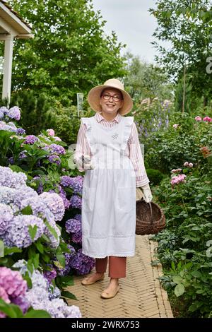 bella donna matura e gioiosa con cappello e occhiali che posa accanto all'ortensia e guarda la macchina fotografica Foto Stock