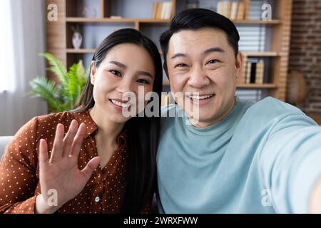 Una allegra coppia asiatica condivide un momento di gioia mentre si fa un selfie in un ambiente accogliente, trasudando felicità e solidarietà. Foto Stock