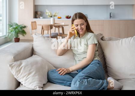 Una donna felice sorridente parla con uno smartphone seduto comodamente sul divano. Una donna positiva fa una chiamata al cellulare Foto Stock
