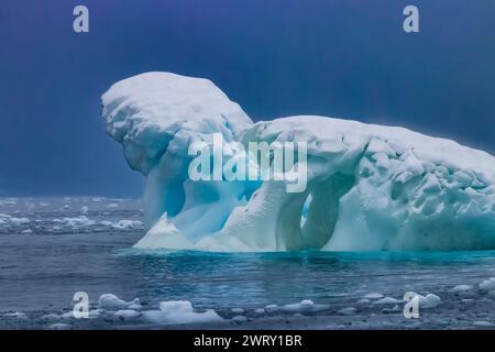 Iceberg scolpito vicino alla penisola antartica. Forma arrotondata e fori nel ghiaccio blu. Riflessione sull'acqua, il ghiaccio scorre in primo piano. Foto Stock