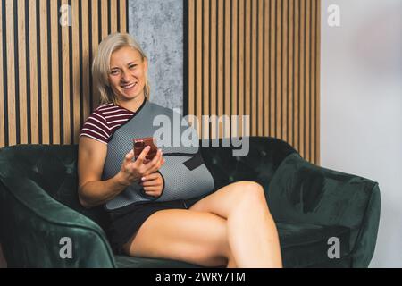 Sorridente donna caucasica con una fionda sulla mano rotta seduta sul divano e con un telefono in mano. Foto di alta qualità Foto Stock