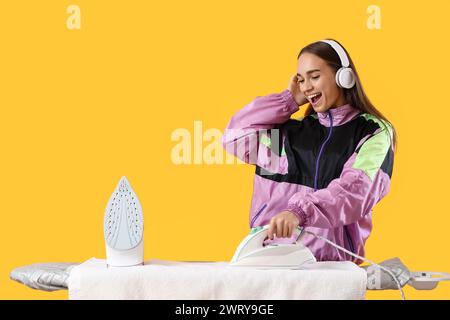 Bella giovane donna con le cuffie che stirano i vestiti mentre ascolta la musica su sfondo giallo Foto Stock