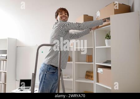 Giovane venditore online su scala a cavalletto che prende il pacco dallo scaffale del magazzino Foto Stock