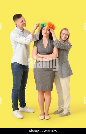 Uomini d'affari che mettono la parrucca da clown sul loro collega su sfondo giallo. Festa del Fool's Day di aprile Foto Stock