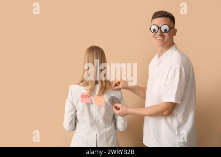 Giovane che attacca il pesce di carta sulla schiena del suo collega su sfondo beige. Festa del Fool's Day di aprile Foto Stock