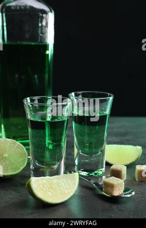 Absinthe in bicchieri da shot, cucchiaio, cubetti di zucchero di canna e lime su un tavolo testurizzato grigio, primo piano. Bevanda alcolica Foto Stock