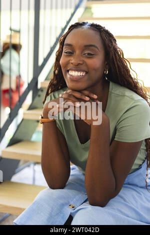 Una giovane donna afroamericana è seduta sulle scale a casa, sorridendo calorosamente Foto Stock