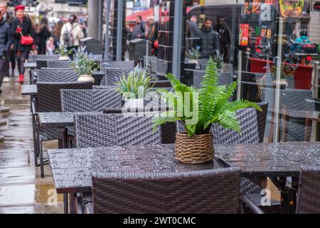 Caffetteria con marciapiede bagnata dalla pioggia con tavoli e sedie bagnati, piante in vaso, pedoni sullo sfondo sfocato. Centro di Londra, Inghilterra. Foto Stock