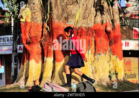 Una ragazza in uniforme rossa passa davanti a un albero ampiamente dipinto di colore rosso, Pokhara Valley, Pokhara, Nepal Foto Stock