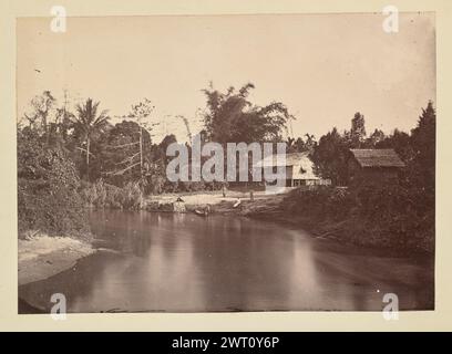 Casa sulle rive di un fiume. Sconosciuto, fotografo anni '1850-'1890 veduta di due edifici con tetto di paglia seduti ad una curva in un fiume. Gli edifici, uno una casa principale e uno un capannone più piccolo, si trovano su palafitte in cima a una sponda del fiume in pendenza, dove due uomini si trovano di fronte all'acqua, e una donna siede tra di loro. Un terzo uomo si trova a vita profonda nel fiume, tenendo sul lato di una canoa. Proprio accanto alla canoa, legata sulla riva del fiume, c'è una seconda barca con un baldacchino di paglia. Foto Stock