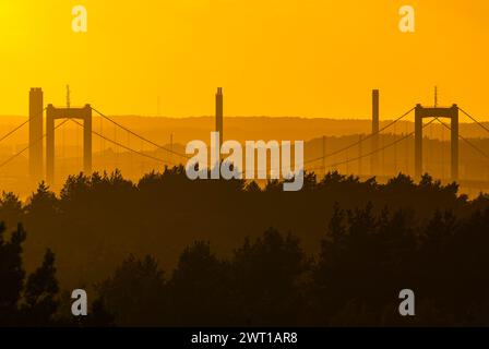 Il sole tramonta all'orizzonte, proiettando un caldo bagliore su un ponte nascosto in lontananza. Il cielo è dipinto con sfumature di arancio e rosa come il giorno Foto Stock